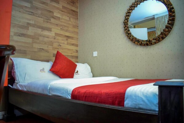 A hotel room with a wooden floor, a bed with white bedding and a red blanket, a round mirror with a brick frame, and a wall socket.