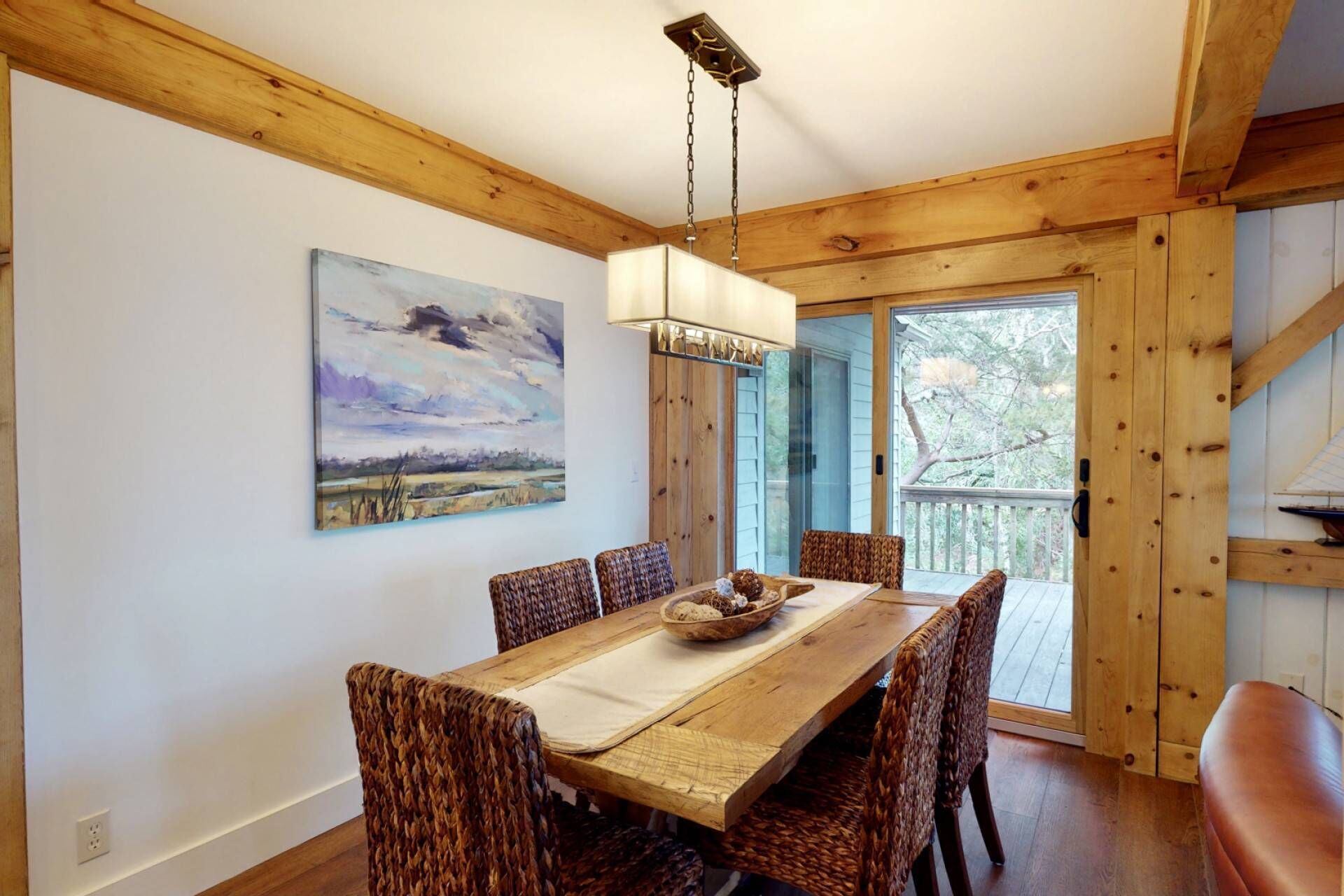 A dining area with a wooden table, wicker chairs, a painting on the wall, and a view of the outdoors through a glass door.