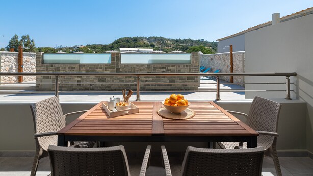 A wooden table with wicker chairs on a balcony with a stone wall and glass windows, overlooking a hillside. - Villa Divina PoolFront Suites (Kremasti)