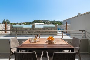 A wooden table with wicker chairs on a balcony with a stone wall and glass windows, overlooking a hillside. - Villa Divina PoolFront Suites (Kremasti)