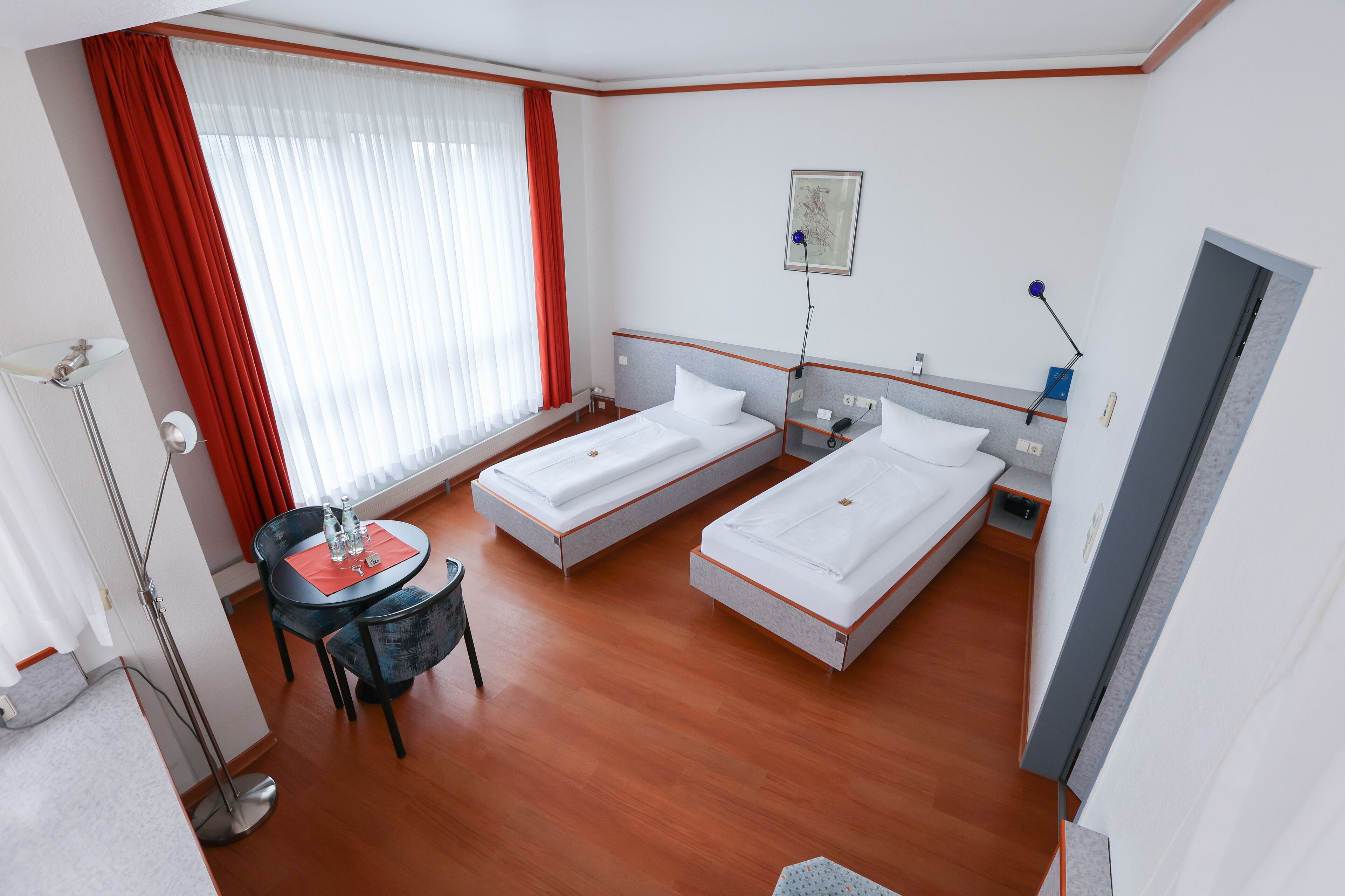 Quarto de hotel com duas camas de solteiro, uma escrivaninha com cadeira, uma mesinha com cadeira, um abajur e uma janela com cortinas vermelhas.