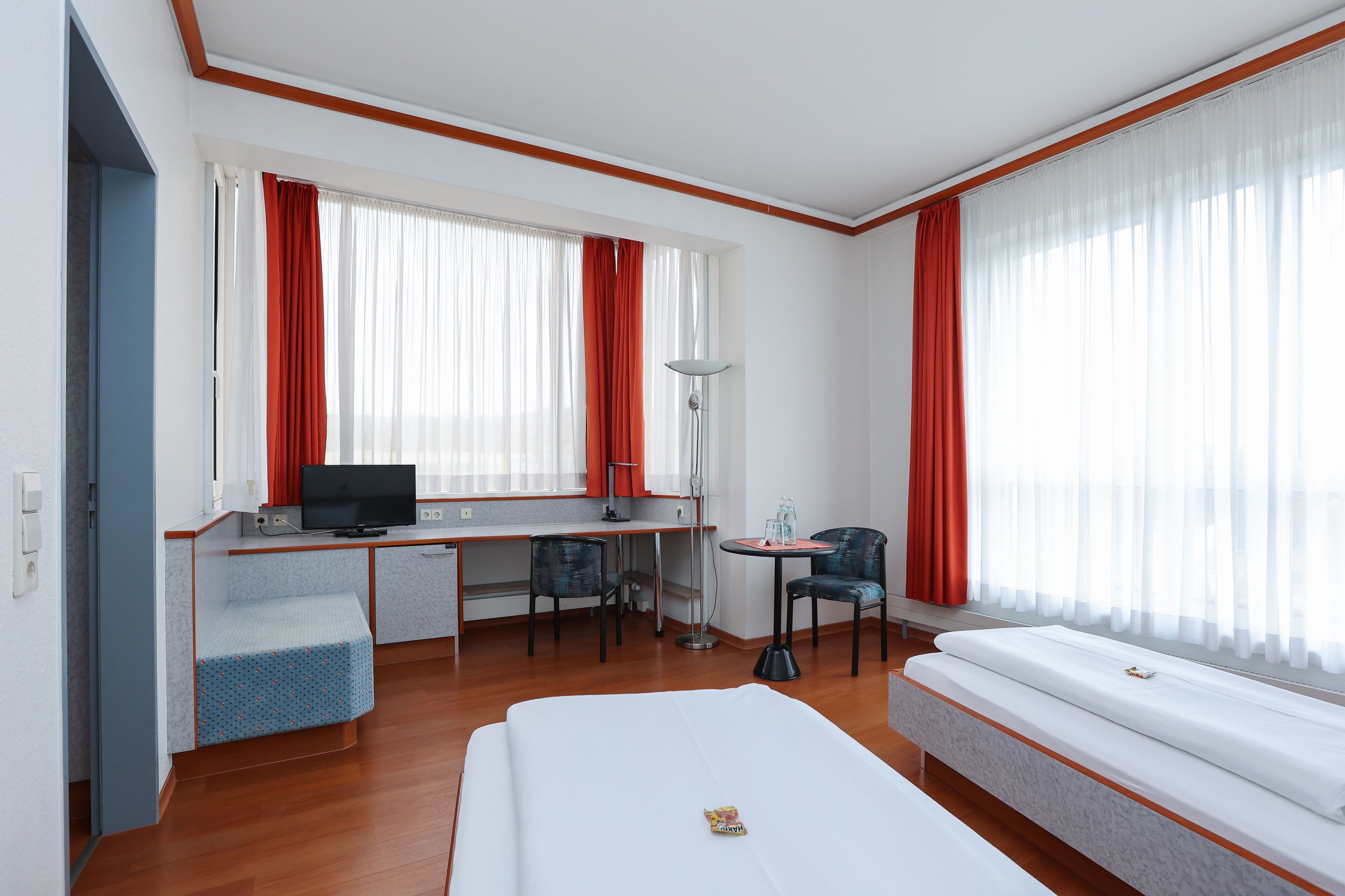 A hotel room with a bed, a desk with a computer, a chair, a small table with a lamp, and large windows with curtains.
