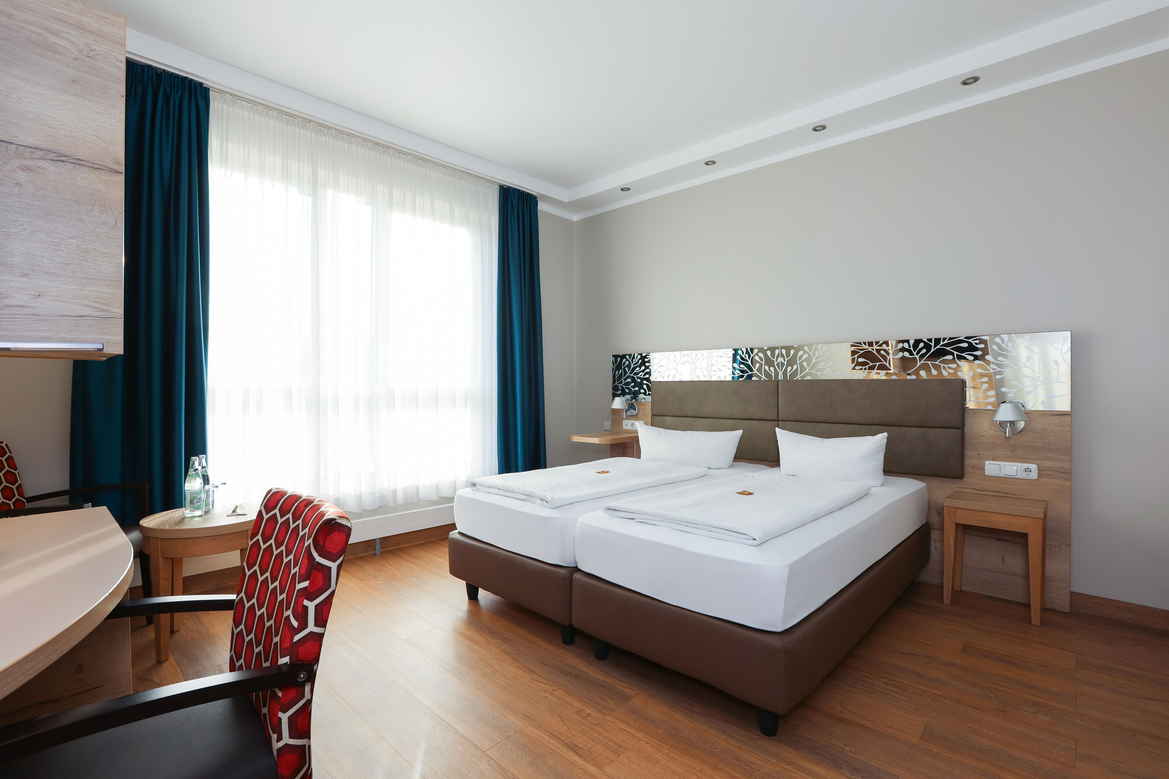 Quarto de hotel com cama de casal, escrivaninha com cadeira, mesinha de cabeceira com abajur e janela grande com cortinas.