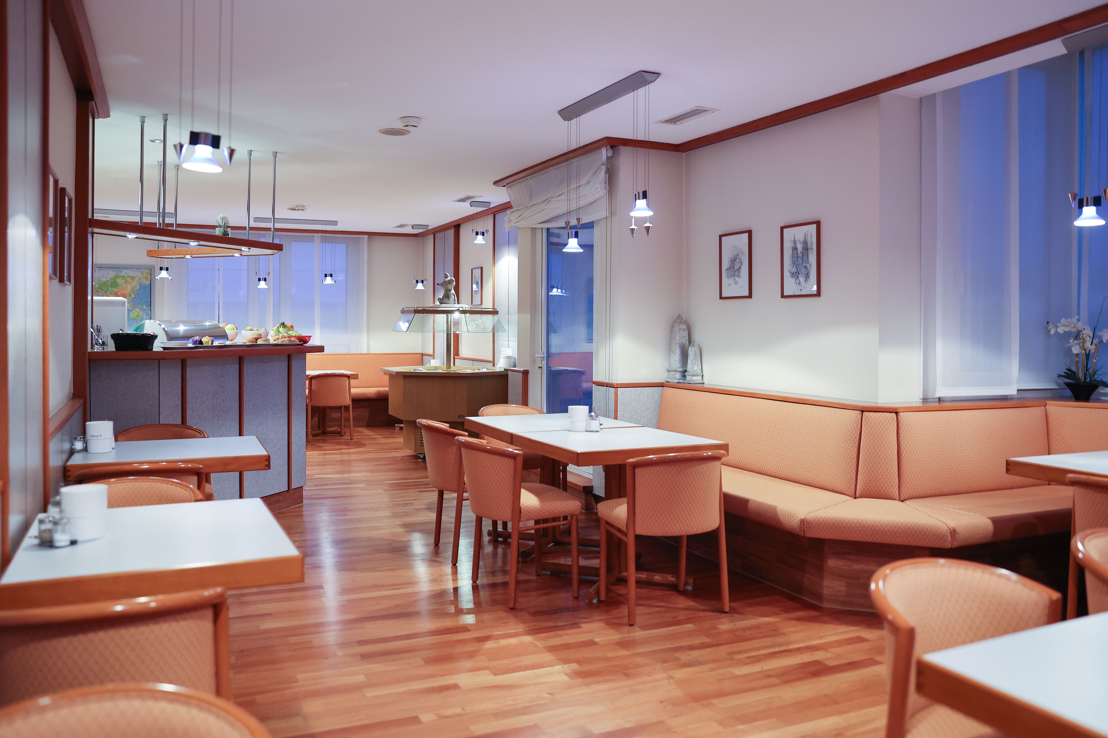 Un comedor moderno con suelo de madera, mesas blancas y asientos con cojines naranja.