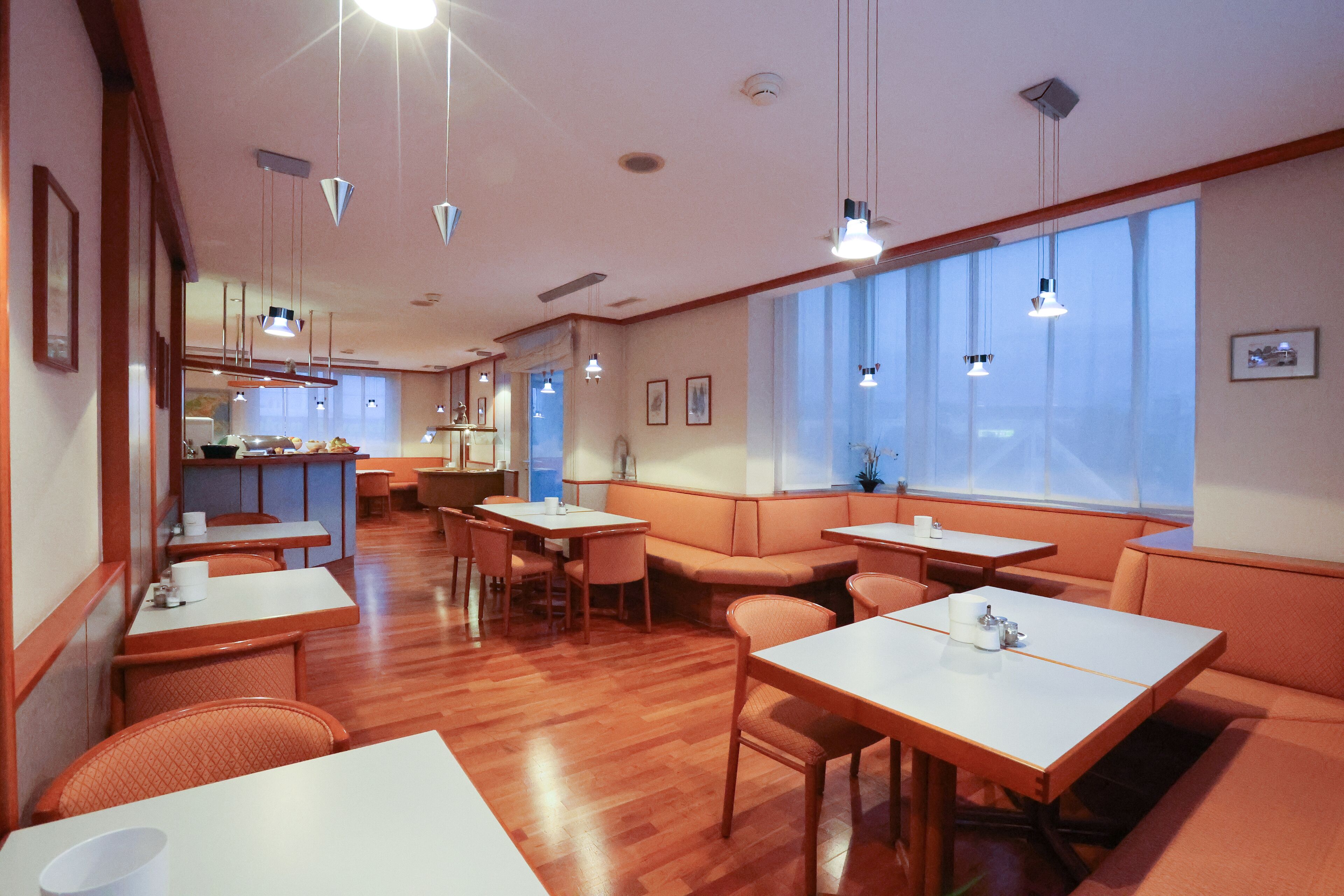 A restaurant with wooden flooring, white tables, orange booths, and pendant lighting.