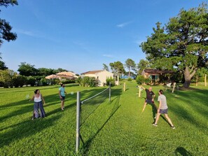 Un espacio exterior con césped, una red de bádminton, árboles y casas al fondo.