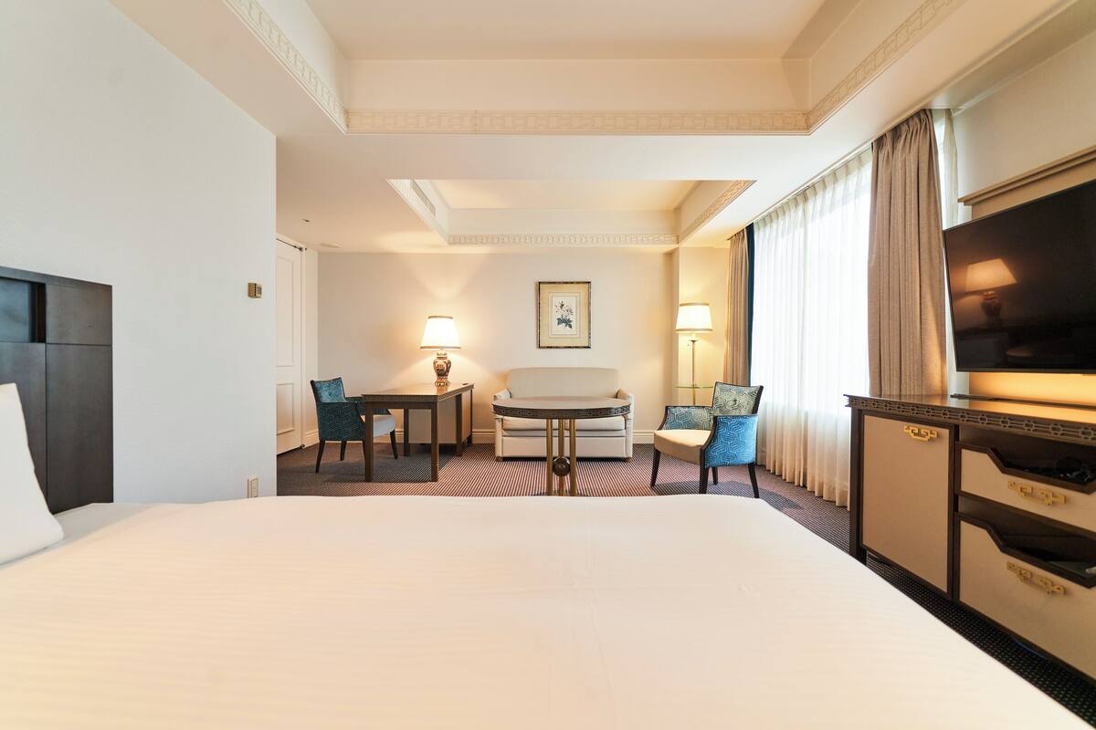 Ein Hotelzimmer mit einem großen Bett, einem kleinen runden Tisch, zwei Stühlen, einer Couch, einem Fernseher und einem Schreibtisch mit Lampen.