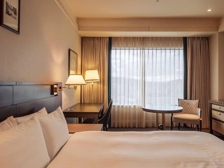 Ett hotellrum med en stor säng, ett skrivbord med en lampa, en stol, ett litet bord och utsikt över staden genom stora fönster.