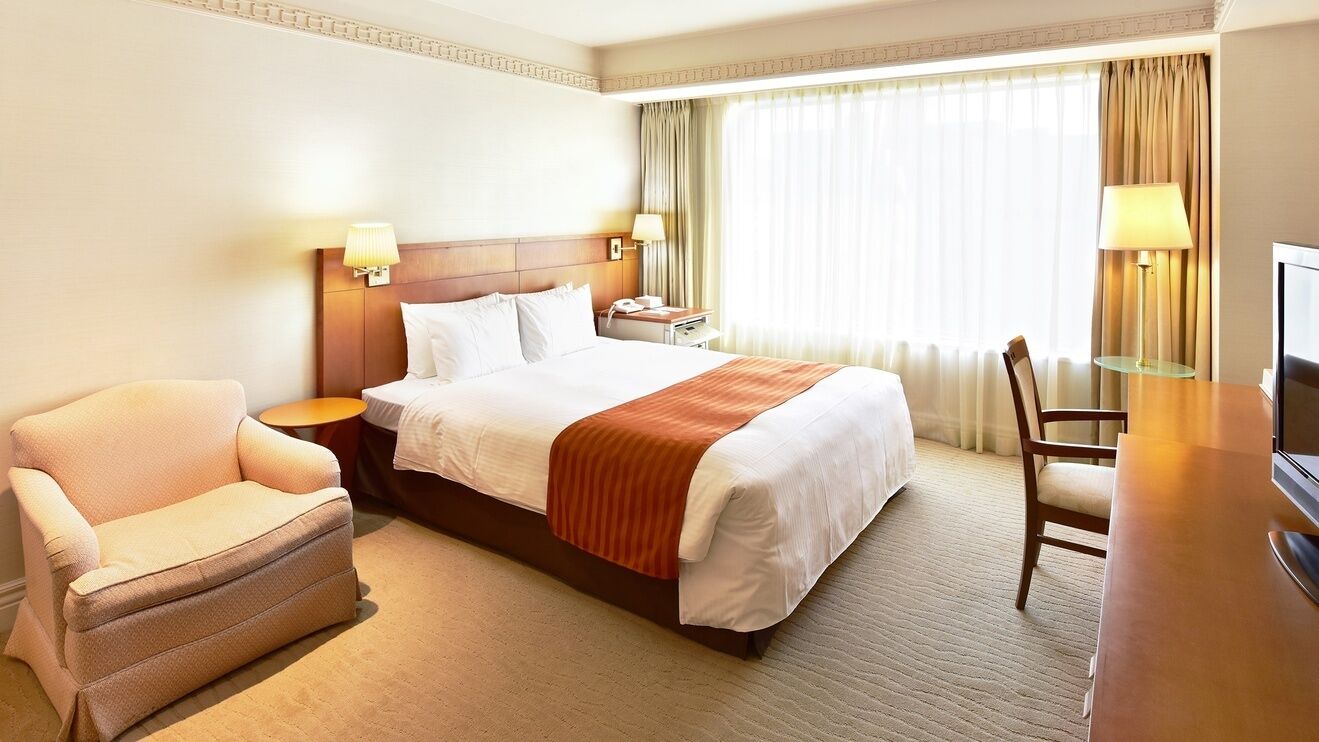 Ein Hotelzimmer mit Bett, Sessel, Schreibtisch, Fernseher und einem Fenster mit Vorhängen.