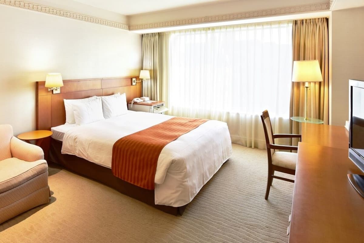 Ein Hotelzimmer mit Bett, Sessel, Schreibtisch, Fernseher und einem Fenster mit Vorhängen.