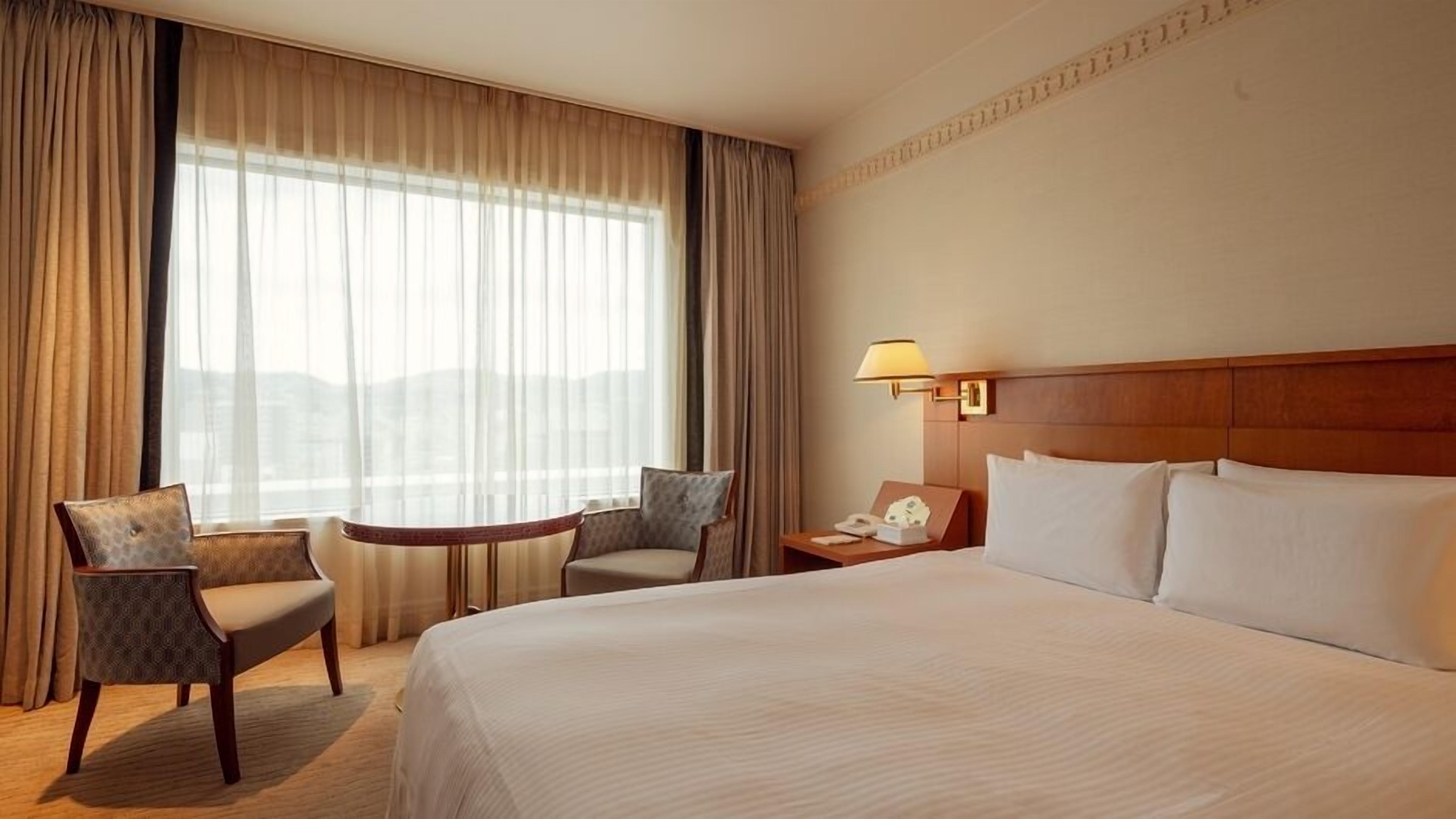 Ein Hotelzimmer mit einem großen Bett, zwei Sesseln, einem kleinen Tisch, einem Nachttisch mit Lampe und Blick auf die Stadt durch transparente Vorhänge.