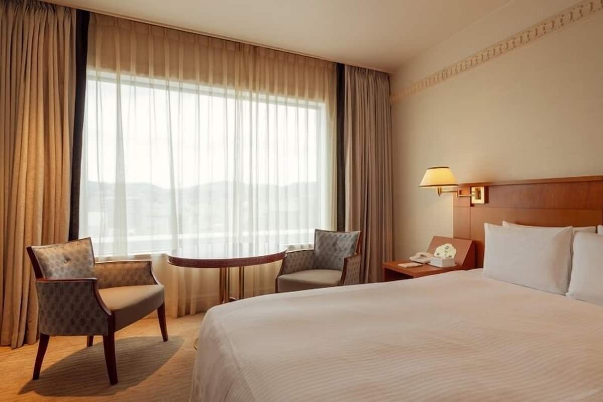 Ein Hotelzimmer mit einem großen Bett, zwei Sesseln, einem kleinen Tisch, einem Nachttisch mit Lampe und Blick auf die Stadt durch transparente Vorhänge.