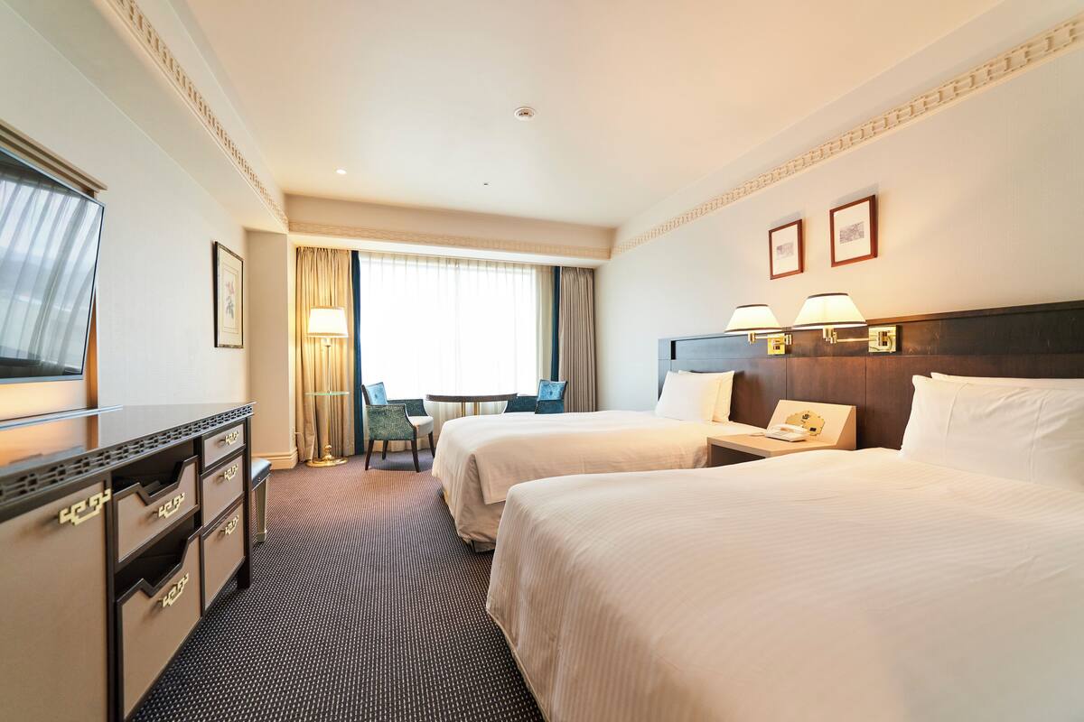 Ein Hotelzimmer mit zwei Betten, einem Schreibtisch, einem Stuhl, einem Fernseher und einem Fenster mit Vorhängen.