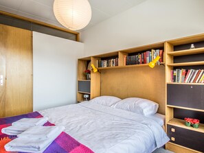 Een net opgemaakt bed met witte lakens, een kleurrijke deken en een stapje opgevouwen handdoeken. Er staan houten boekenplanken met boeken en decoratieve objecten.