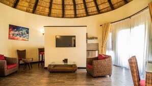 A spacious living area with a thatched ceiling, wicker furniture, a flat-screen TV, and a large window with curtains. - Hunters Paradise Cottages (Bungoma)