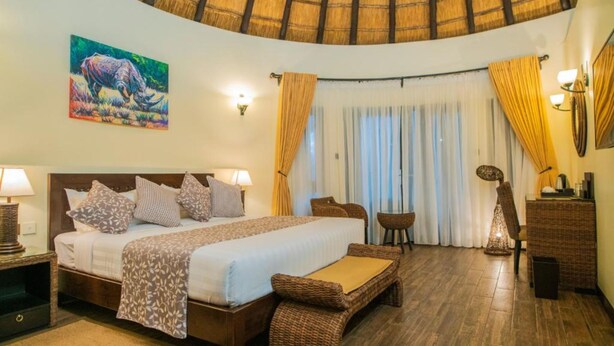 A hotel room with a bed, a nightstand, a wicker chair, a small table, and a lamp. - Hunters Paradise Cottages (Bungoma)