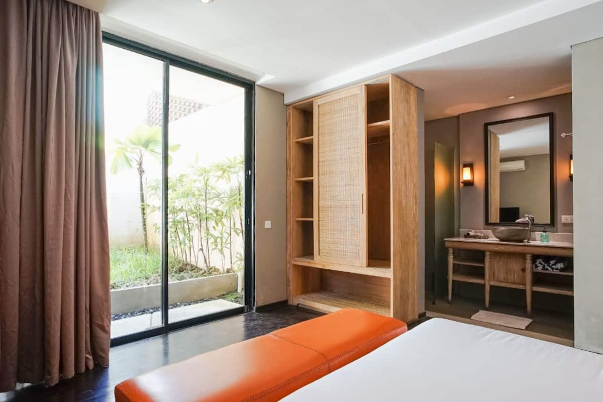 Une chambre d’hôtel moderne dotée d’une grande porte-fenêtre coulissante donnant sur un jardin, d’une armoire en bois, d’une salle de bain avec un lavabo et un miroir, et d’un banc orange.