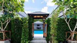 A pathway leading to a pool area with a covered seating area and a view of a building in the background. - Lespalm Taraburi Pool Villa (Si Sunthon)