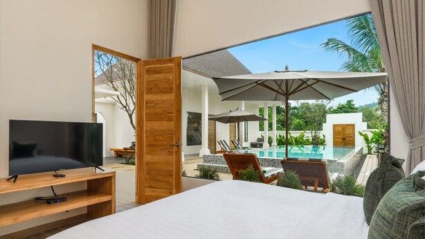 A bedroom with a TV, a large bed, and a view of an outdoor pool area. - Lespalm Taraburi Pool Villa (Si Sunthon)