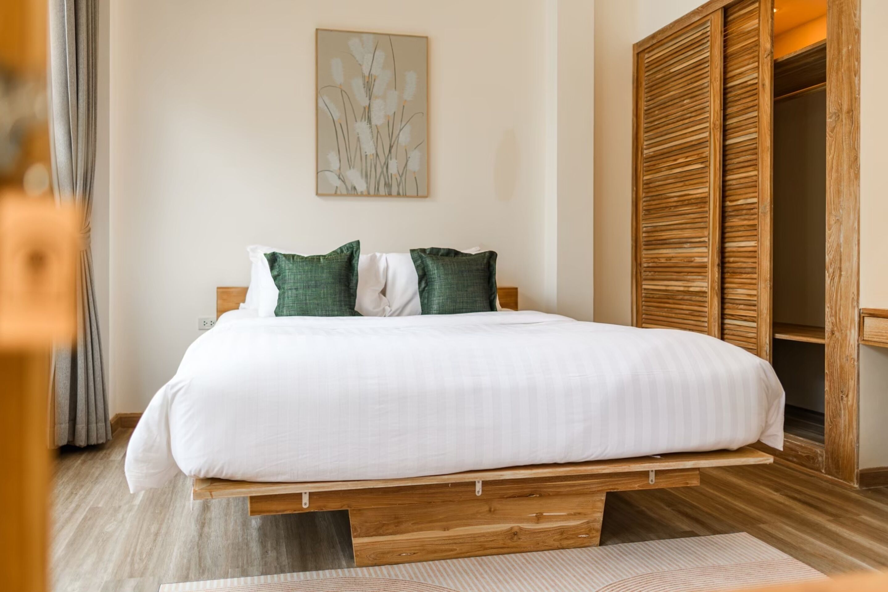 A bedroom with a wooden platform bed, green and white pillows, a wall-mounted artwork, and a wooden wardrobe.