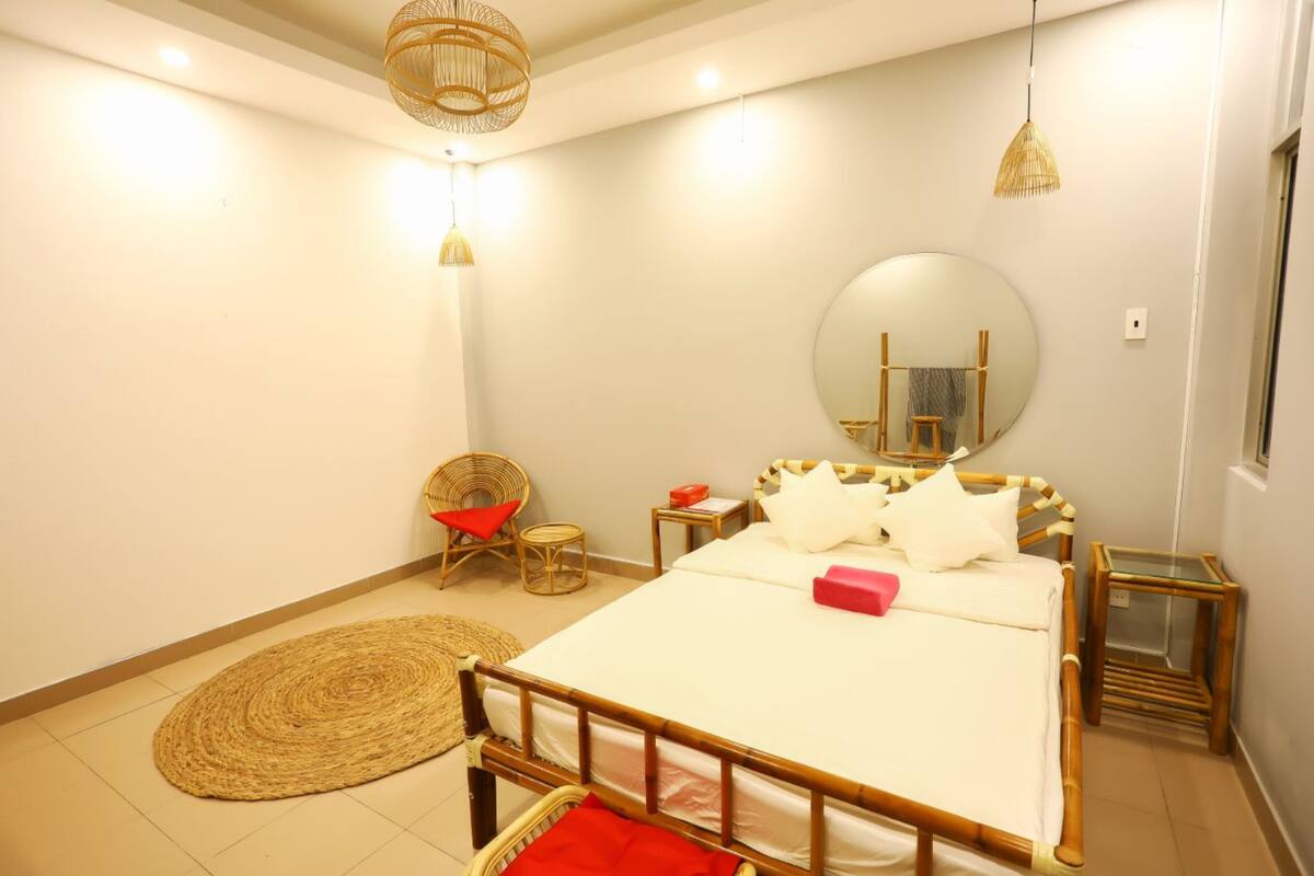 Um quarto com uma cama, um espelho redondo, uma cadeira de vime e uma mesa de cabeceira.