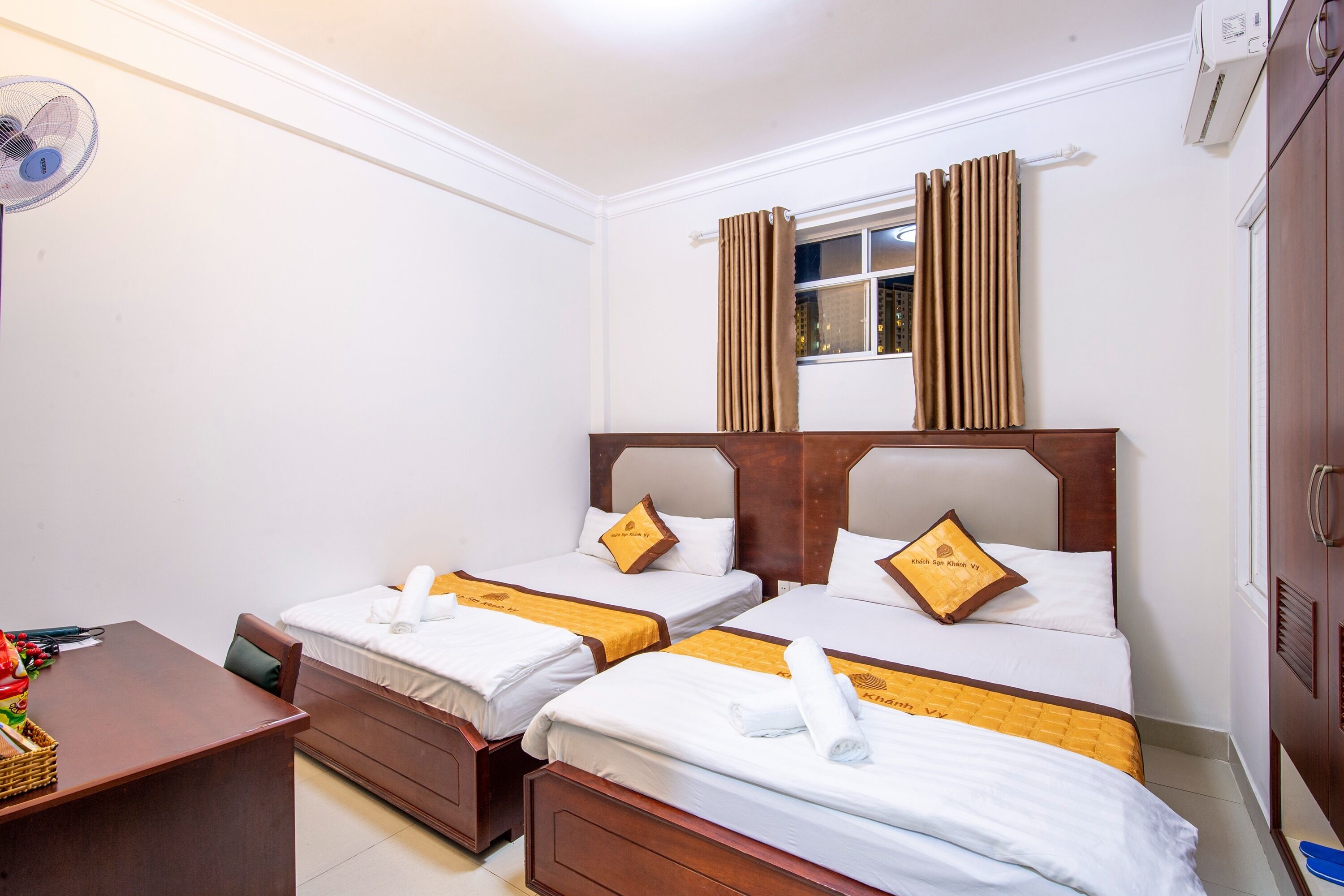 A hotel room with two single beds, a wooden desk, a fan, and a window with curtains.