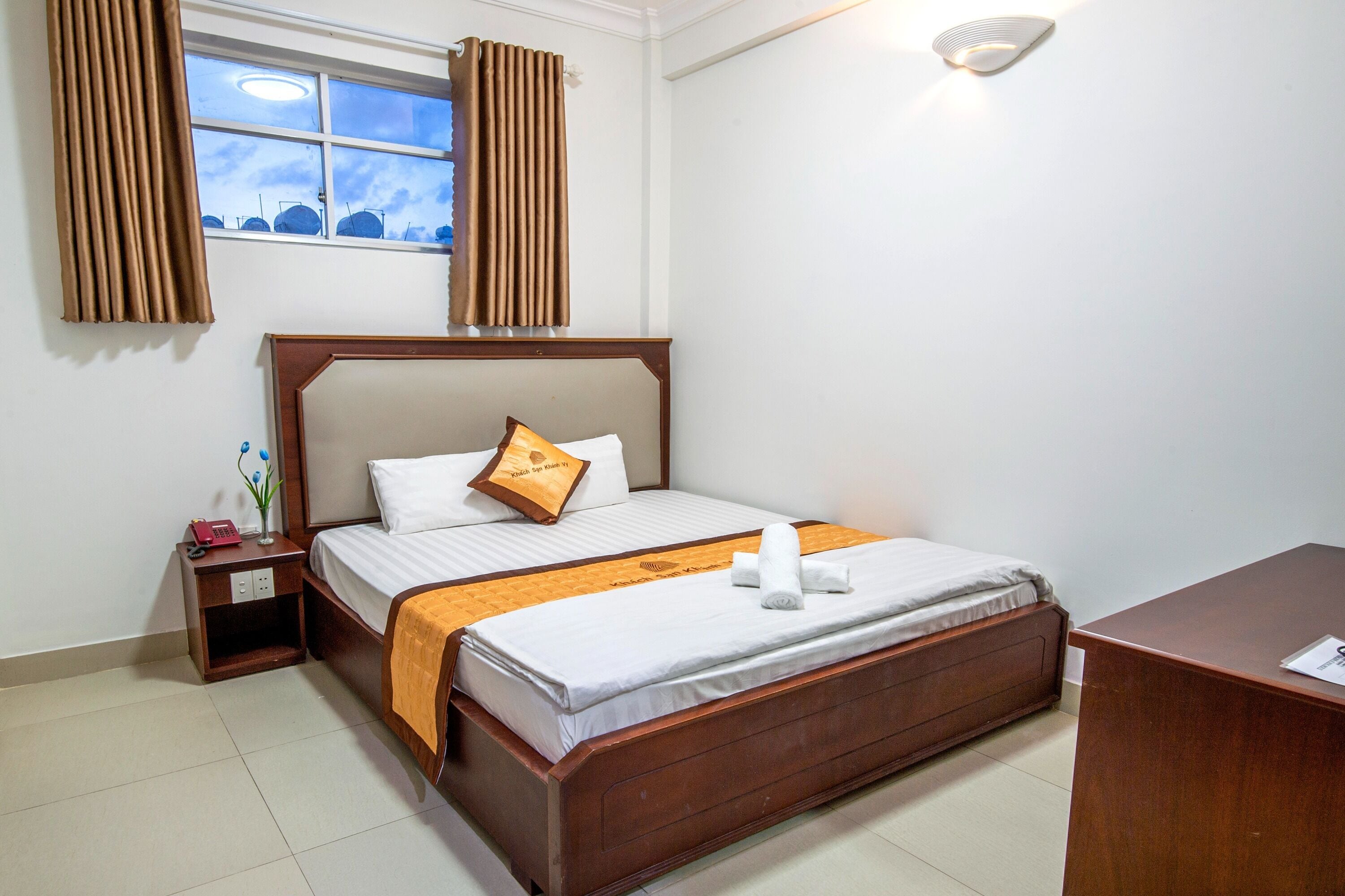 Habitación de hotel con cama de madera, mesita de noche, escritorio y ventana con cortinas.