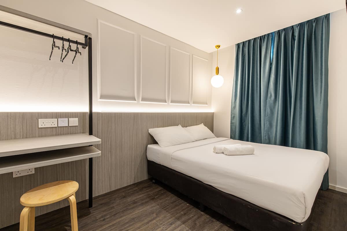 A hotel room with a bed, a desk, a chair, a hanging light fixture, and a window with curtains.