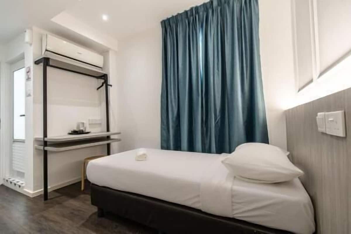 A single bed with a white mattress and pillow, a desk with a chair, a wall-mounted air conditioning unit, and a window with blue curtains.