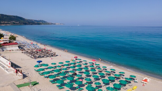 A beach with rows of green umbrellas and sun loungers, a clear blue sea, and a hilly landscape in the background. - Residence Emmesse Trilocal 3 Rooms (Zambrone)