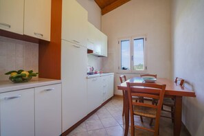 Cozinha com armários brancos, uma mesa de jantar de madeira e cadeiras. Há uma janela com vista para o verde lá fora.
