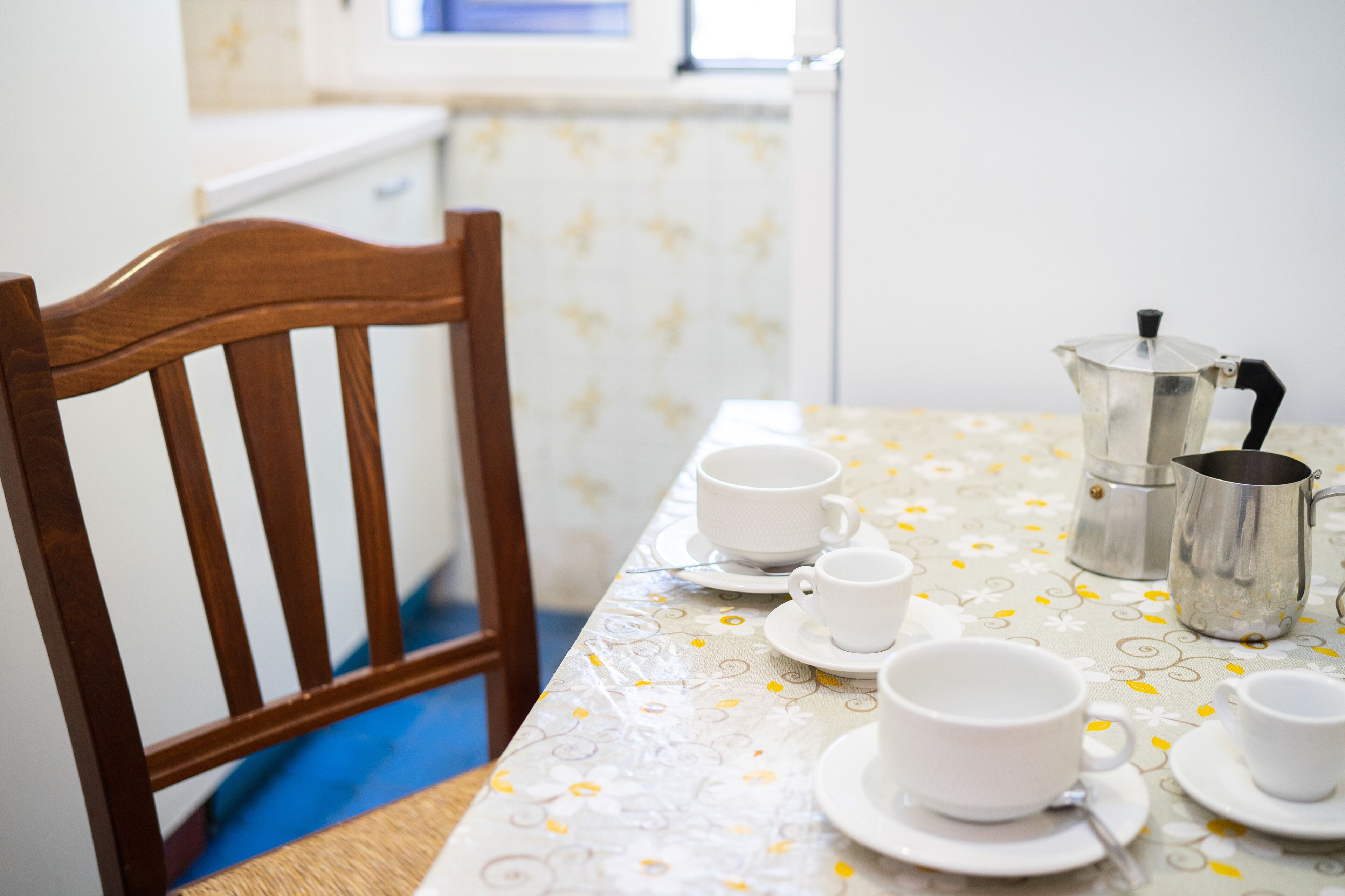 A dining table with a floral tablecloth, a metal coffee pot, and several white cups and saucers.