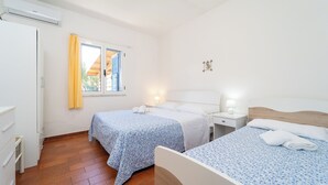 Et hotelværelse med to senge, et vindue med gule gardiner, en hvid aircondition og et lille natbord med lampe.