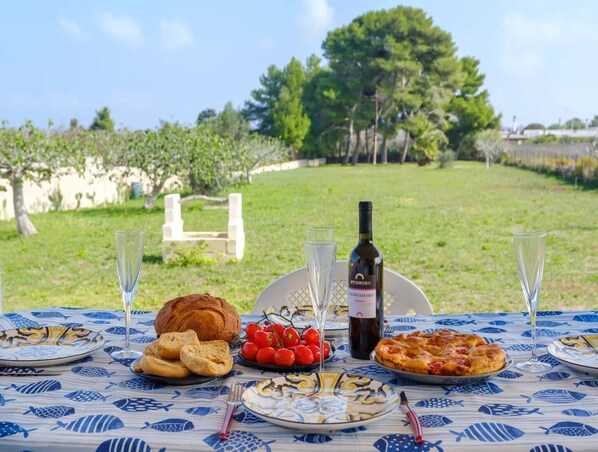 A table set outdoors with a bottle of wine, glasses, bread, tomatoes, and a pizza.
