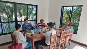 Un grupo de personas sentadas alrededor de una mesa de madera en una habitación con ventanas amplias, algunas sosteniendo platos y cubiertos, lo que sugiere un ambiente para comer.