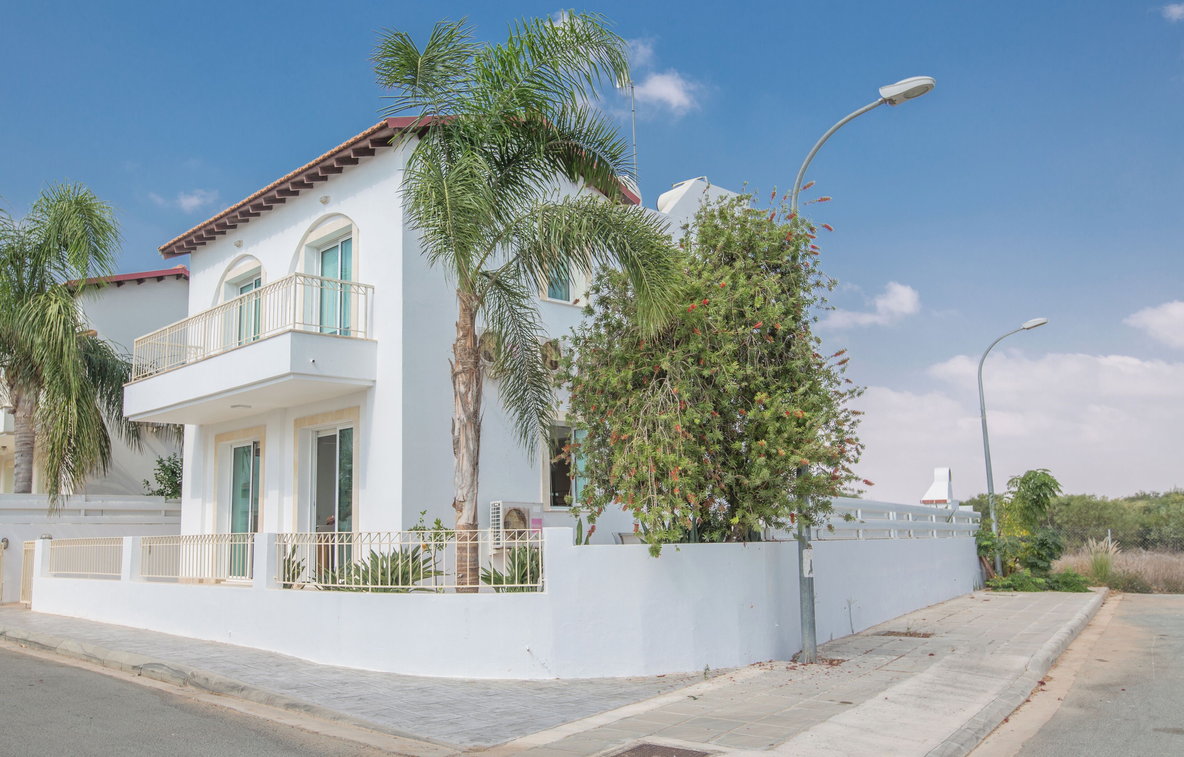 A two-story white building with a balcony, surrounded by palm trees and a white fence.