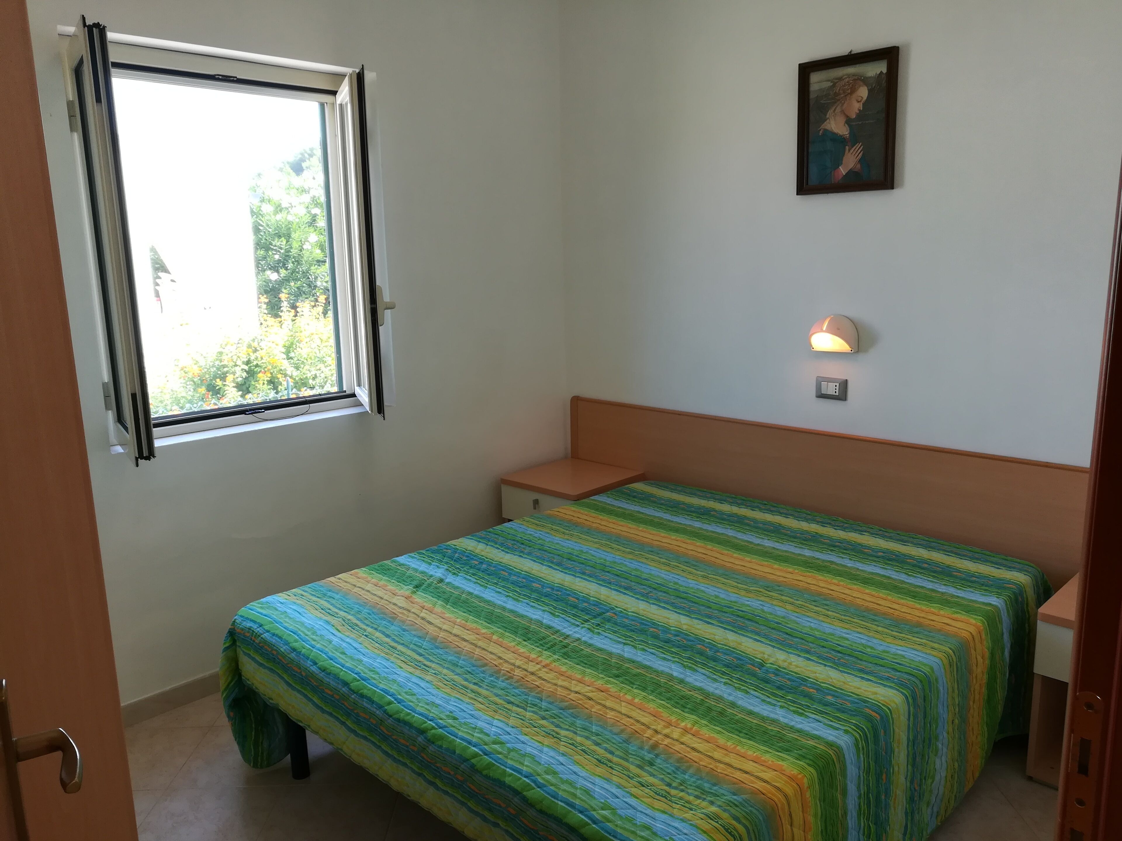A small, simple room with a bed, a nightstand, a wall-mounted lamp, and a window with an open curtain.