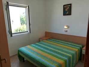 Een kleine, eenvoudige kamer met een bed, nachtkastje, wandlamp en raam met open gordijn.