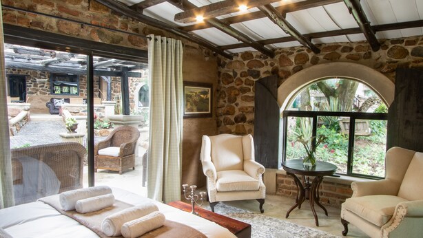 A room with a bed, two armchairs, a table, and a view of an outdoor patio.