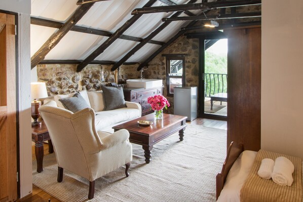 Una habitación acogedora con pared de piedra, vigas de madera, un sofá blanco, un sillón de mimbre, una mesita de madera y un lavamanos pequeño.