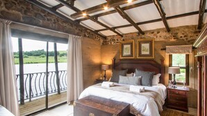 A bedroom with a large bed, a wooden headboard, two bedside tables with lamps, a nightstand, and a view of a green landscape through a balcony.