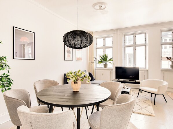 A modern living room with a round dining table, comfortable chairs, a black woven light fixture, a flat-screen TV, and large windows with white frames. - City 2- Cosy 1 Bdr Apt In the Center of Copenhagen (Copenhagen)