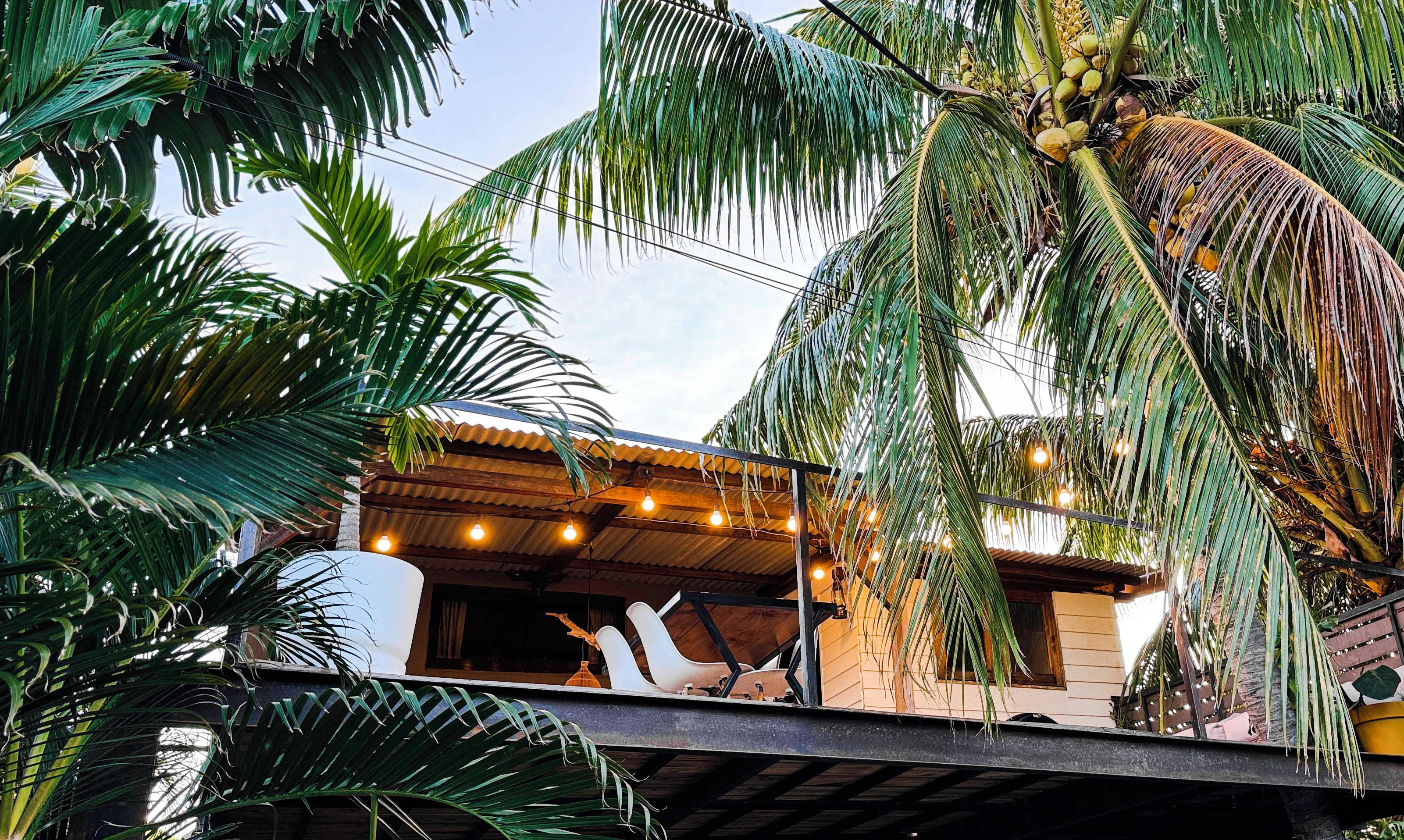 A tropical setting with a building featuring a corrugated metal roof and a balcony with hanging lights.