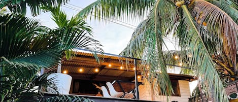 A tropical setting with a building featuring a corrugated metal roof and a balcony with hanging lights.