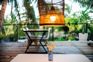 A table with a clear glass vase containing dried flowers, a wicker hanging light fixture, and a wooden deck with chairs and potted plants in the background. - Ti Bout Paradi Ti Zil (Tamarin)