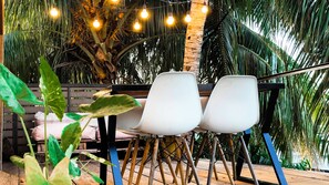 A patio area with a table and two white chairs, surrounded by lush greenery and string lights.