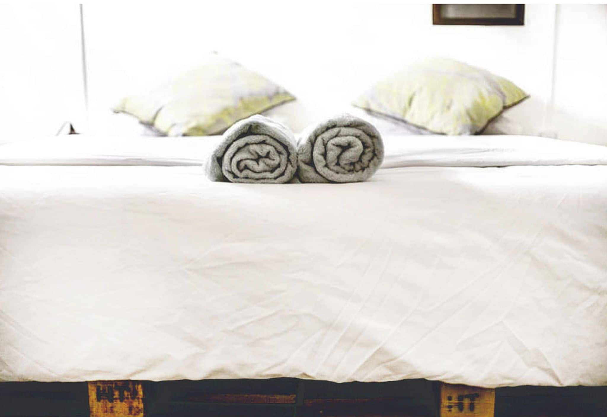 A neatly made bed with rolled towels in the center, a white bedspread, and two pillows on the bed.