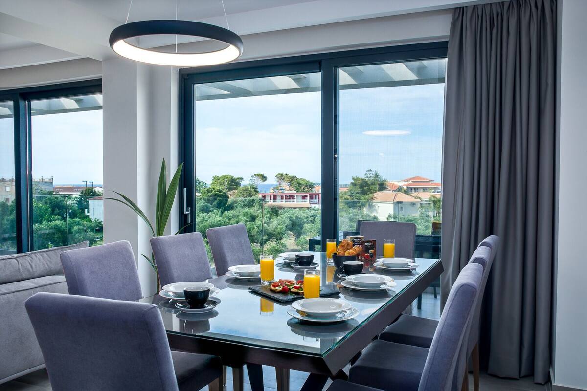 A modern dining area with a glass table, grey chairs, and a city view through large windows.