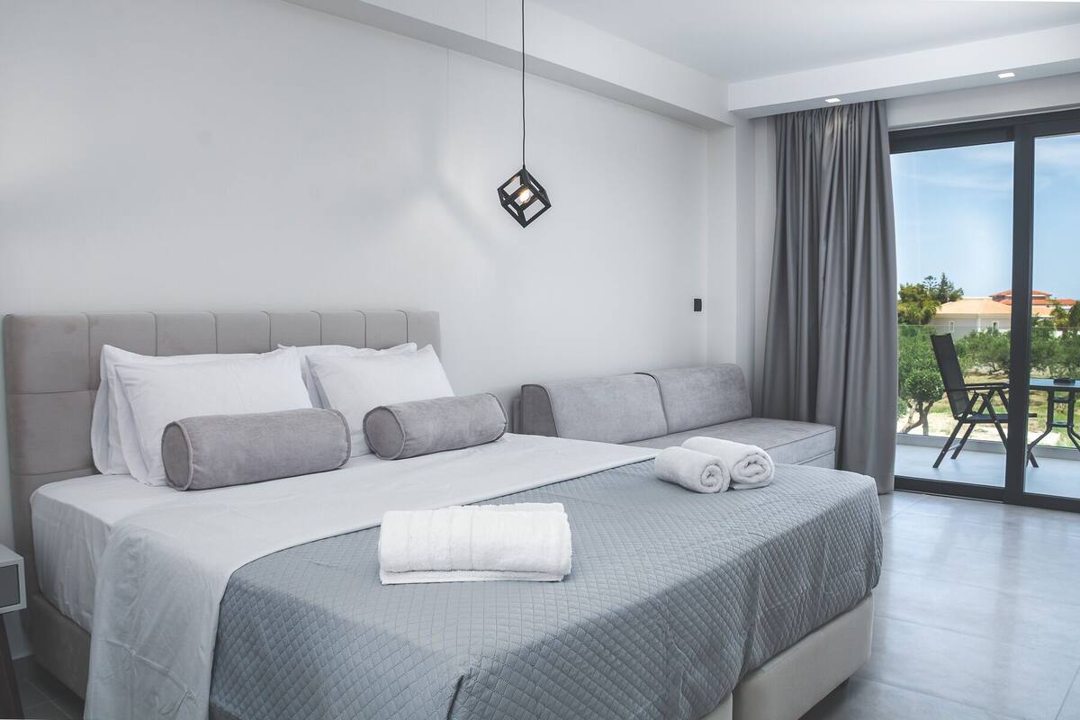 A modern bedroom with a large bed, grey bedding, and a view of the outdoors through a sliding glass door.