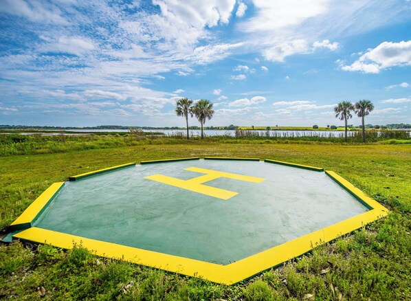 A large, flat, green and yellow marked area with a central yellow 'H' symbol, surrounded by grass and palm trees, with a body of water and a cloudy sky in the background.